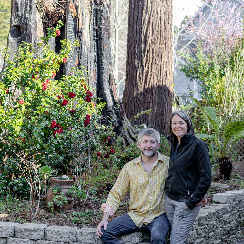 Andrew and Kristin smile in front of the flowers, fern and redwood trees in their backyard.