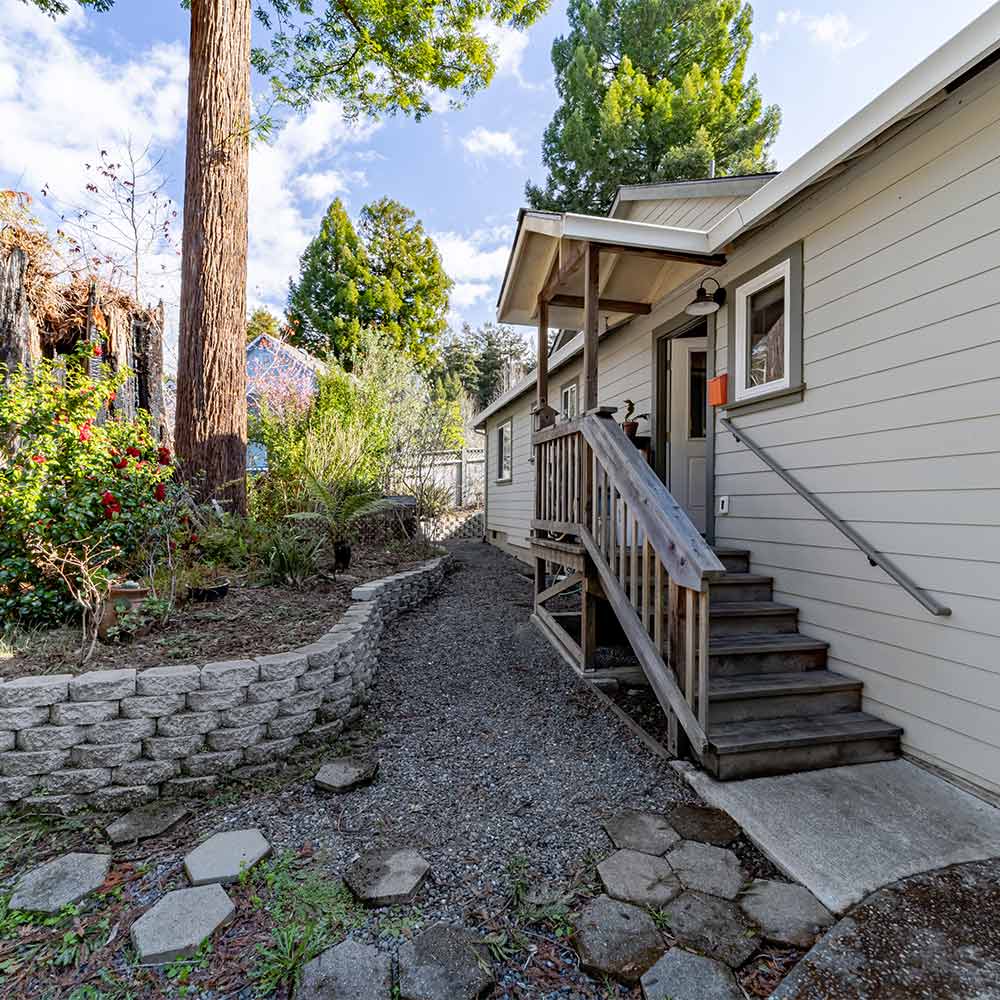 gravel walkway leading along residence, landscaped backyard with ornamental plants and redwood trees, and the porch entrance to the junior ADU.