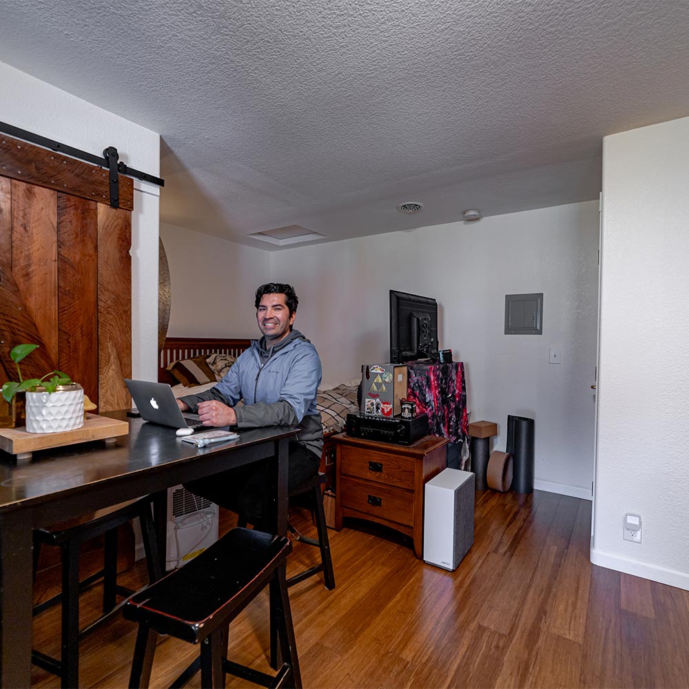 Tenant who lives in junior ADU smiling at dining table with bed and television in the background.