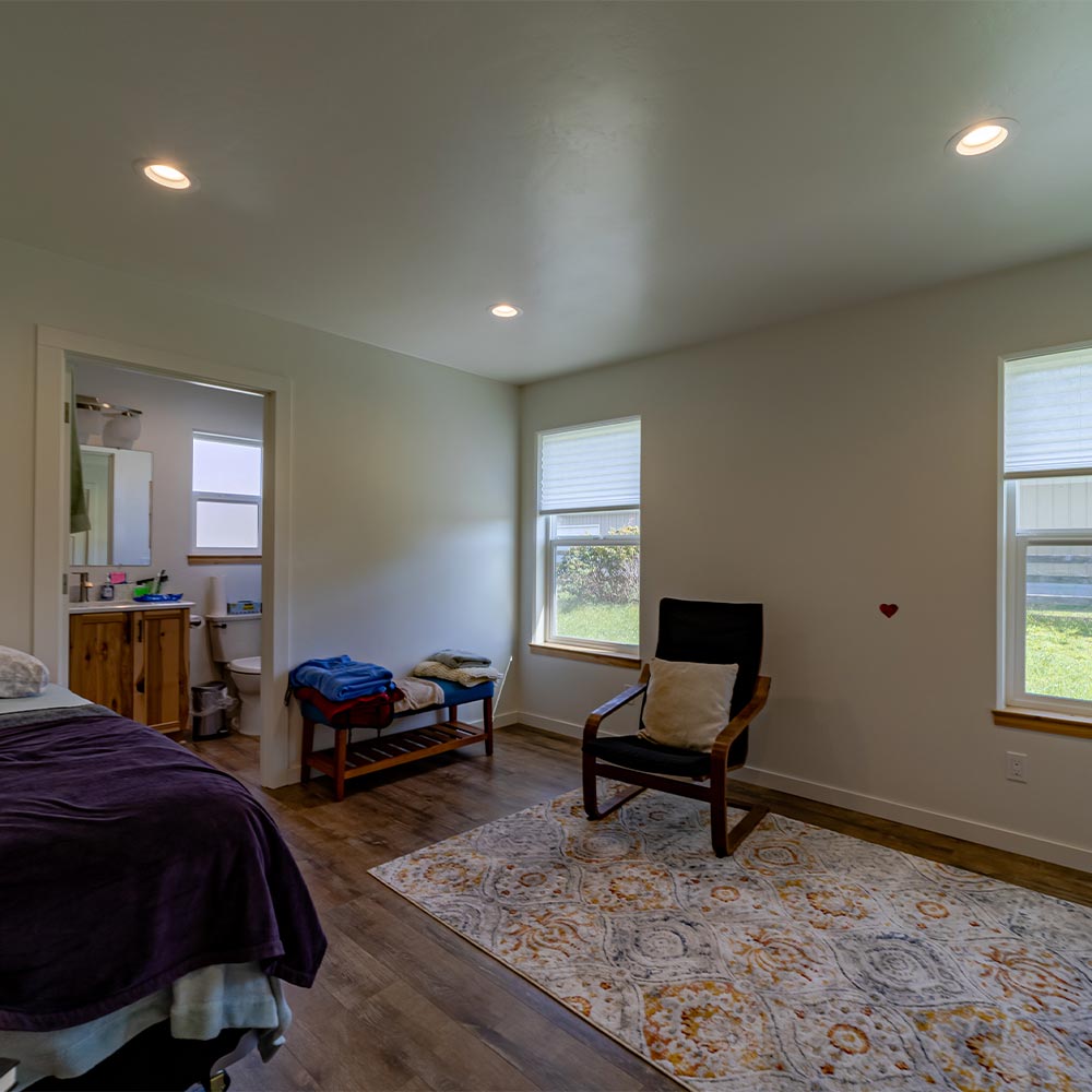 Interior of the second bedroom with plenty of natural light, a sitting chair on a patterned rug and door open into the bathroom.