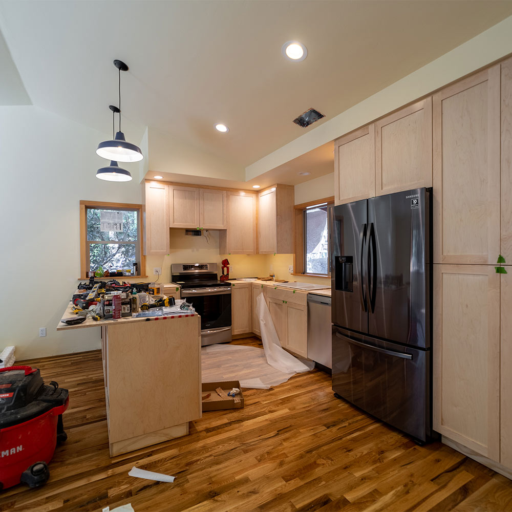 Modern kitchen with two windows, hanging lights, fridge, dishwasher, stove, and wooden cabinets. Island covered with construction materials.